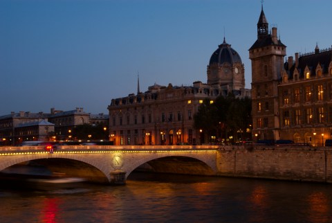 Image of softly illuminated buildings next to a river flowing under a stone bridge.