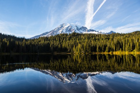 Image of snowy mountain behind a green forest reflected in a still lake under a sky streaked with clouds