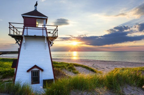 A white lighthouse next to a grassy beach as the sun sets over the ocean.