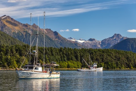 Two small fishing boats in a calm harbor with trees and mountains rising up in the distance on a sunny day.
