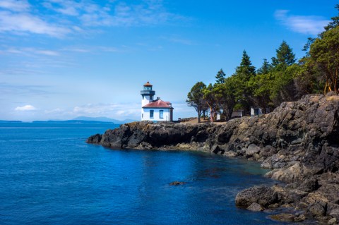 Image of a small white lighthouse on a rocky forested promontory jutting into deep blue ocean.
