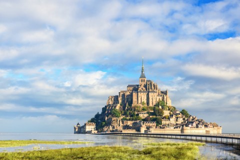 A gothic, stone abbey on an island rising from marshy water and green grass under cloudy blue sky
